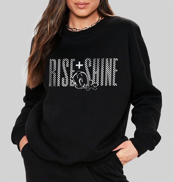 Front view of a woman in a black sweatshirt featuring a vibrant 'Rise and Shine' design with a stylized checkered graphic and uplifting text.