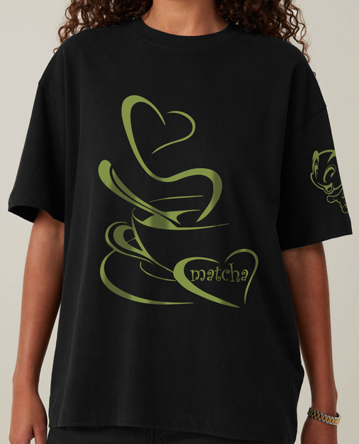 A woman wearing a black t-shirt with a hand-illustrated heart-shaped matcha latte design on the front, featuring creamy green tones and latte art details.