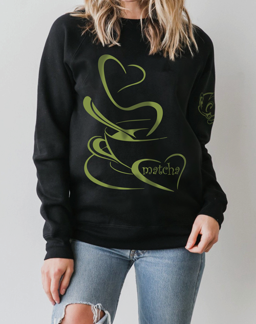 A woman with blonde hair wearing a black sweatshirt with a hand-illustrated heart-shaped matcha latte design on the front, featuring creamy green tones and latte art details.